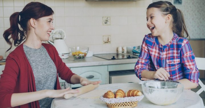 Young mother rolling dough and talking to her little cute daughter while cooking in the kitchen on weekend. Family, food, home and people concept